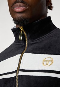 Svart og hvit zip-up fleecejakke med gullglidelås, med et hvitt bånd og gullbrodert logo på brystet. Myk tekstur.