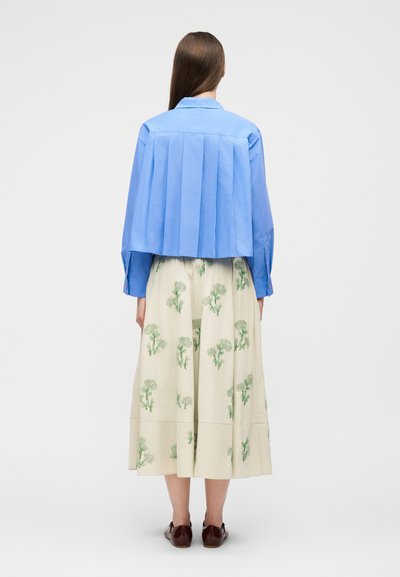 Γυναίκα που στέκεται πλάτη φορώντας πλισέ κοντομάνικο γαλάζιο πουκάμισο, μπεζ φούστα με πράσινα λουλουδένια σχέδια και καφέ παπούτσια σε λευκό φόντο.