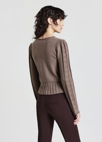 Brauner Strickpullover mit geripptem Saum, Puffärmeln mit Kabeldetails und Rundhalsausschnitt. Getragen mit dunkelbraunen Hosen.