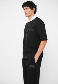 Νεαρός άντρας φορώντας μαύρο κοντομάνικο πουκάμισο Armani Exchange και αντίστοιχες μαύρες παντελόνες με λευκή ρίγα, στέκεται με το ένα χέρι στην τσέπη.