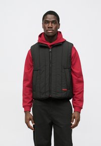 Gilet nero trapuntato con cerniera frontale, indossato sopra una felpa con cappuccio rossa. Presenta due tasche frontali e un logo rosso. Pantaloni neri completano l'outfit.