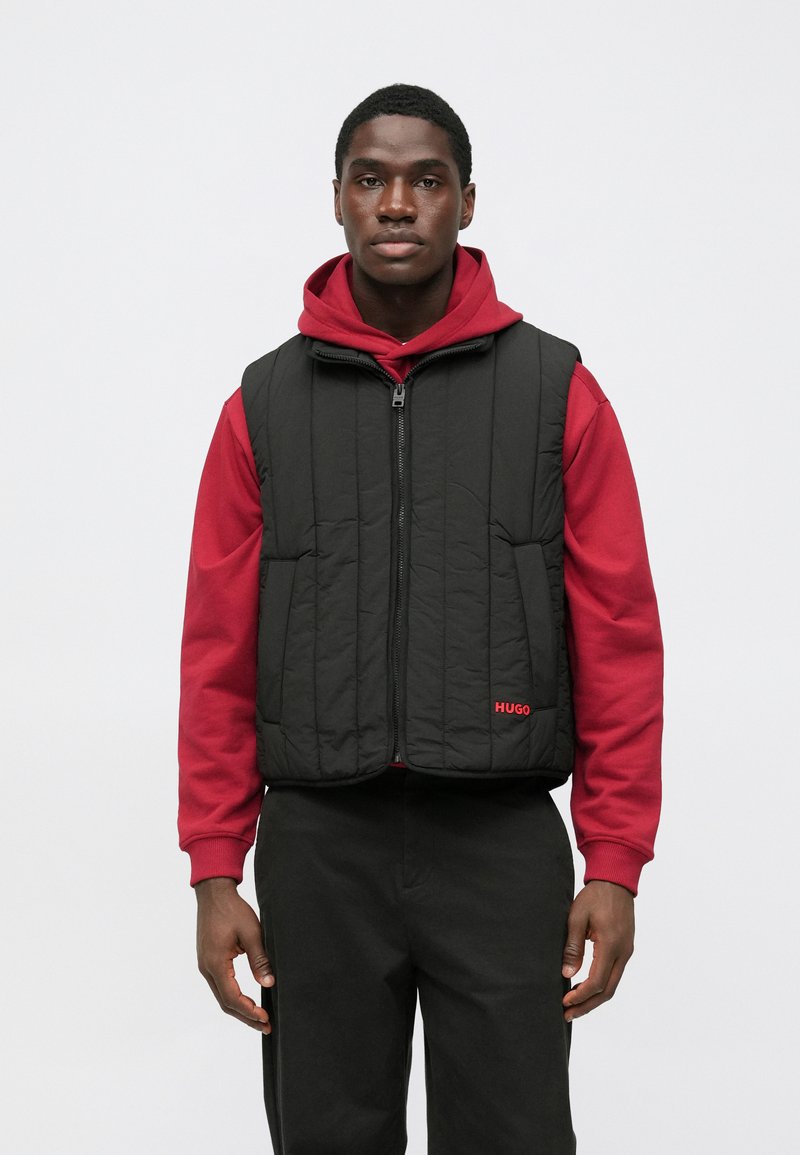 Gilet nero trapuntato con cerniera frontale, indossato sopra una felpa con cappuccio rossa. Presenta due tasche frontali e un logo rosso. Pantaloni neri completano l'outfit.