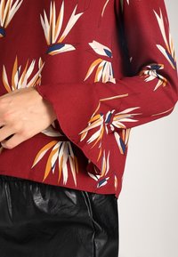 Rød bluse med blomsterdesign i hvid, blå og orange. Har flagrende ærmer og let, glat stof. Bæres sammen med en sort struktureret nederdel.