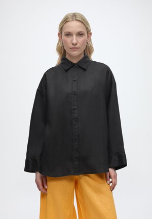 Femme aux longs cheveux blonds portant une chemise noire ample à boutons et un pantalon large jaune, debout devant un fond blanc uni.