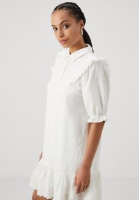 Vit klänning med krage, spetsdetaljer, puffärm och volangkant. Tyget har ett subtilt strukturerat mönster.