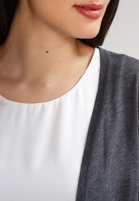 Cardigan grigio appoggiato su una blusa bianca. Il tessuto appare morbido e sottile, con una texture liscia e un design a frontale aperto. Dettagli di cucitura sottili.