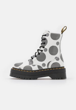 Bota blanca con cordones, con patrones circulares a rayas en blanco y negro, suela negra gruesa y detalle de costura amarilla.