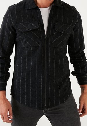 Mann trägt schwarze Sweatjacke mit Reißverschluss, dünnen vertikalen Streifen und zwei Brustklappentaschen über einem weißen T-Shirt.