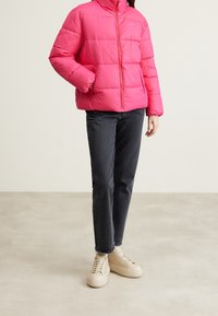 Giacca imbottita rosa con cuciture orizzontali, colletto alto e zip frontale. Abbinata a jeans scuri e scarpe da ginnastica di colore chiaro.