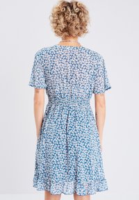 Robe fleurie avec un fond bleu, présentant des motifs de fleurs blanches, des manches courtes et une taille smockée. L'ourlet est légèrement volanté.