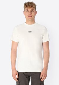 Young man with short curly blond hair wearing a plain white t-shirt with small black text on chest and dark cargo pants.