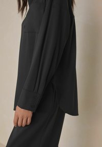 Black long-sleeve shirt made from smooth fabric, featuring a chest pocket, button cuffs, and side slits for ease of movement.