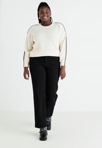 Cremfarbener Rippstrickpullover mit schwarzen Akzenten, kombiniert mit hochgeschnittenen schwarzen weiten Hosen und Ankle Boots. Einfaches Design, strukturiertes Material.