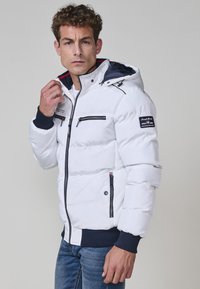 Chaqueta acolchada blanca con capucha, con detalles en azul marino, múltiples bolsillos con cremallera y puños acanalados. Textura suave con diseño acolchado.