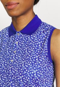 Lila blommig ärmlös polo med en ribbad krage, med mörkblå detaljer och silverfärgade knappar; mjukt tyg med ett detaljerat botaniskt mönster.