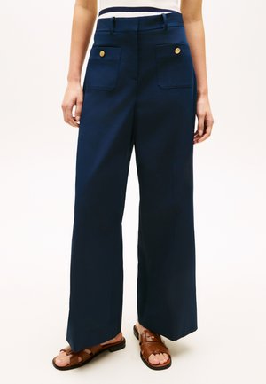 Pantaloni ampi navy a vita alta con due tasche frontali con bottoni dorati, indossati con sandali di pelle marrone.