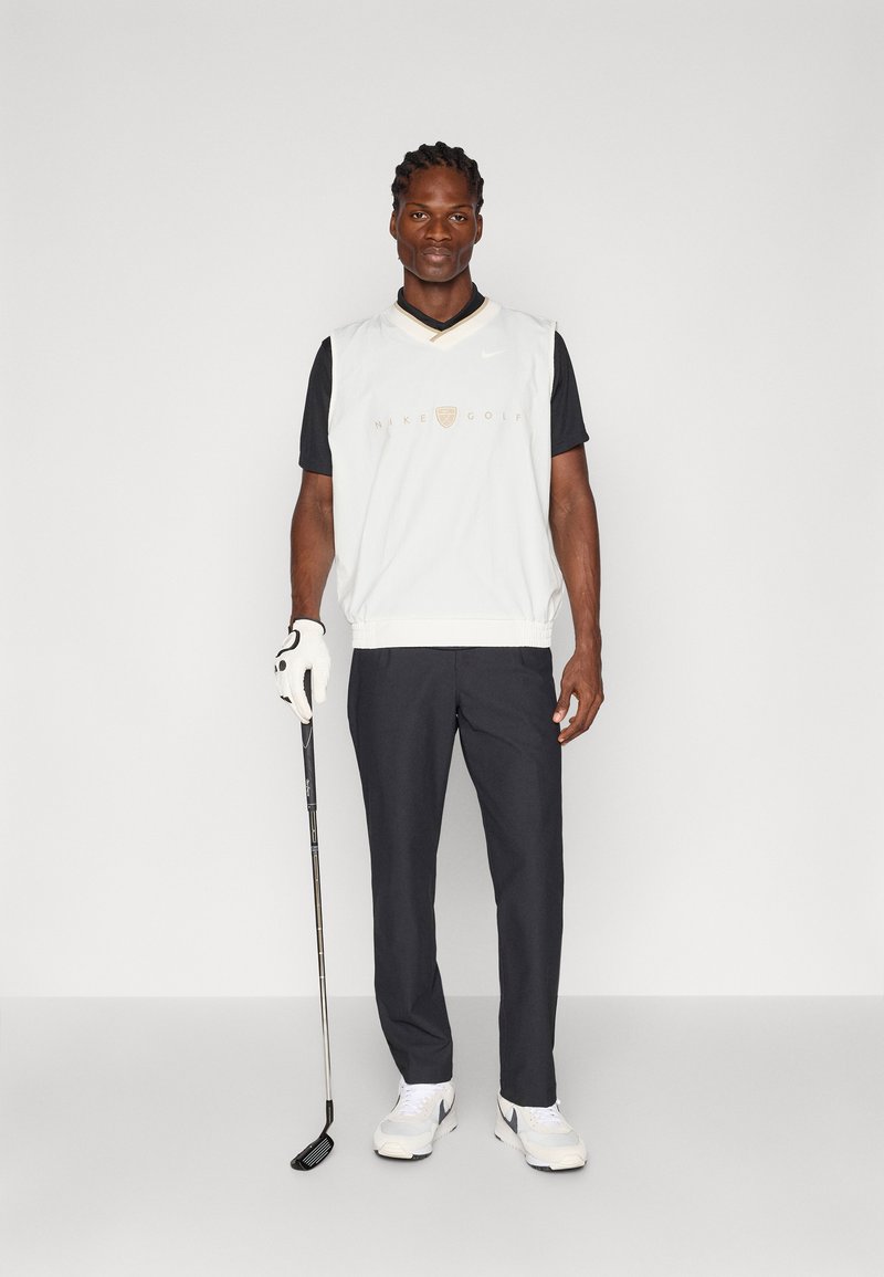 White sleeveless golf top over a black shirt, dark pants, white golf shoes, with a golf club in hand. Features a gold logo at the chest.
