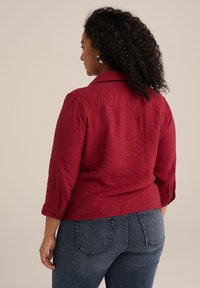 Camicia rossa cropped e testurizzata con colletto e maniche a tre quarti; abbinata a jeans in denim blu scuro con dettagli sulle tasche.