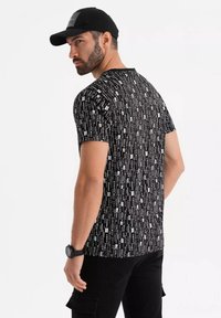 Camiseta negra de manga corta con un patrón de texto blanco vertical. Combinada con una gorra negra lisa y pantalones cargo negros.
