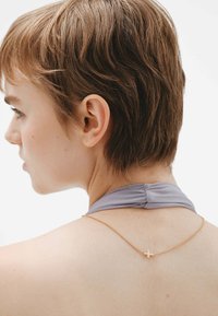 Collar de cadena de oro con un pequeño colgante en forma de cruz, llevado sobre una blusa de tirantes gris, contra un fondo blanco difuminado.
