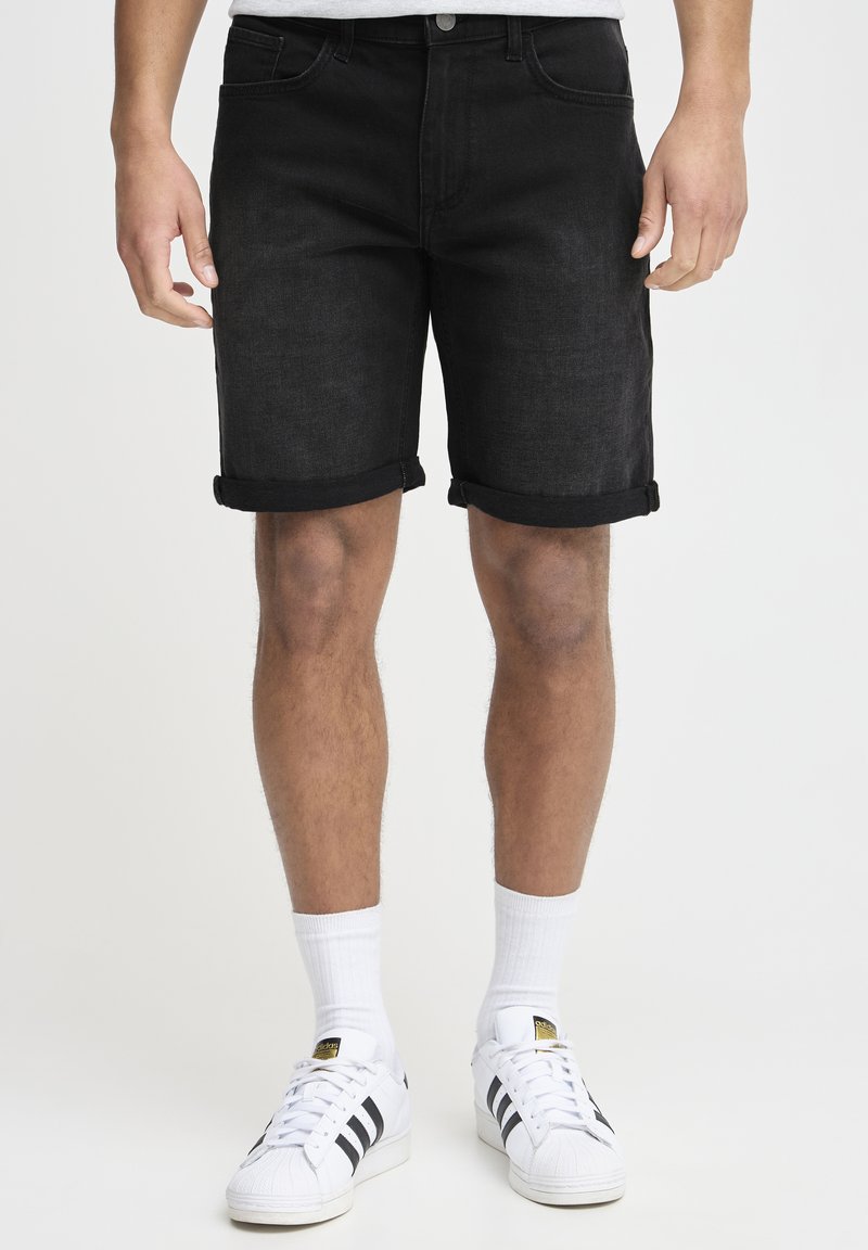 Shorts en denim noir avec un ourlet retroussé, cinq poches et une fermeture à bouton, associés à des chaussettes blanches et des baskets à rayures noires.