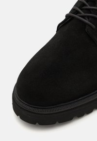 Černá semišová bota s tlustou gumovou podrážkou, zaoblenou špičkou a kontrastním prošitím podél švu. Obsahuje šněrování a texturovaný povrch.