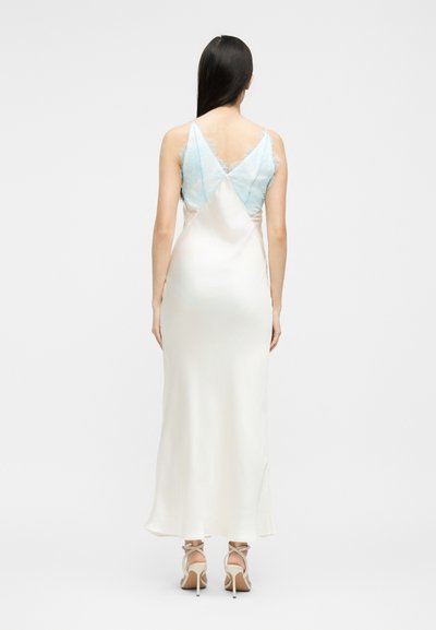 Γυναίκα με μακριά μαύρα μαλλιά που φοράει λευκό σατέν φόρεμα με ανοιχτό μπλε δαντέλα στο πάνω μέρος της πλάτης και λευκές ψηλοτάκουνες σανδάλια με λουράκια.