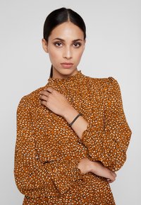 Blusa color ruggine con colletto alto arricciato e maniche lunghe a sbuffo. Presenta una stampa a pois bianca e nera. Tessuto dalla consistenza liscia.