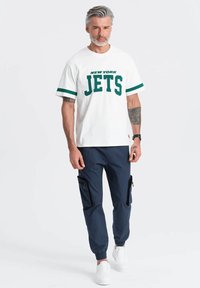 T-shirt blanche avec "NEW YORK JETS" en vert, associée à un pantalon cargo marine avec des poches zippées et des poignets élastiques, portée avec des baskets blanches.
