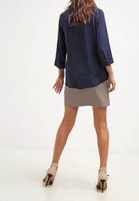 Marinebluse med trekvartærmer, parret med en beige nederdel og nude højhælede sko, præsenteret fra bagsiden.
