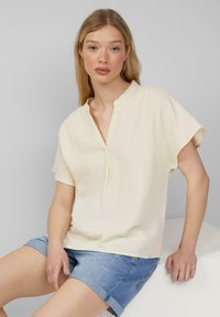 Hellbeige Bluse mit kurzen Ärmeln aus strukturiertem Material, mit halber Knopfleiste und einem weichen, lässigen Schnitt. Kombiniert mit hochgerollten Denim-Shorts.