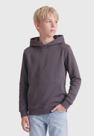 Garçon blond regardant vers la droite portant un sweat à capuche gris foncé et un jean bleu clair, une main dans la poche.