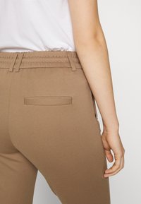 Brune bukser med en glat tekstur, der har elastisk talje og en baglomme. Designet inkluderer en sidelukning.