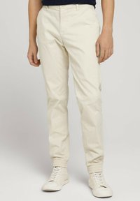 Beżowe spodnie chino o zwężanym kroju, z bocznymi kieszeniami, płaskim przodem i elastycznymi ściągaczami, zestawione z białymi trampkami.