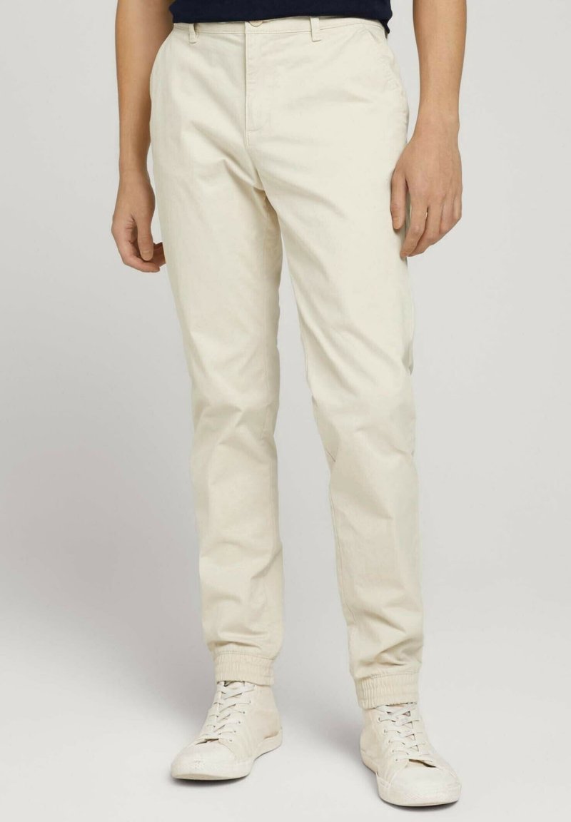 Beżowe spodnie chino o zwężanym kroju, z bocznymi kieszeniami, płaskim przodem i elastycznymi ściągaczami, zestawione z białymi trampkami.