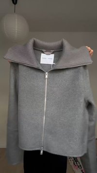 Γκρι ζακέτα με φερμουάρ και φαρδύ ριπ κολάρο, με ετικέτα "Samsoe Samsoe", κρατημένη μπροστά σε απλό φόντο με ένα χαρτόφως πάνω από το κεφάλι.