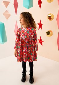 Robe rouge à manches longues avec motifs colorés de fête, jupe évasée et encolure ronde. Associée à des bottines noires et des collants noirs.