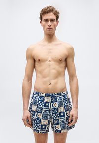 Νεαρός άνδρας στέκεται όρθιος, φοράει μαγιό με μοτίβα σε μπλε, λευκό και καφέ αποχρώσεις που απεικονίζουν ήλιους, φεγγάρια, ψάρια και φοίνικες.