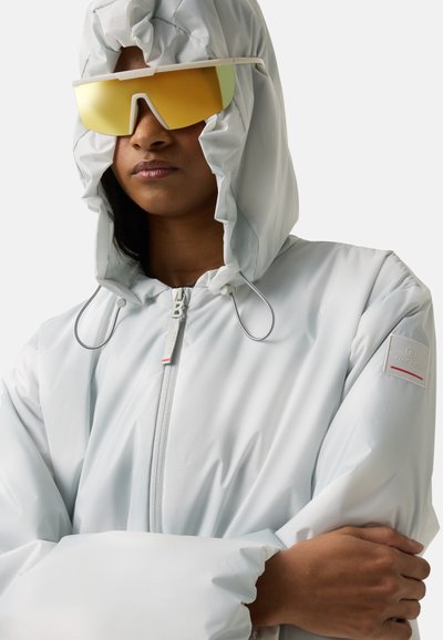 Persoon draagt een wit jack met capuchon en ritssluiting aan de voorkant, met een gele zonnebril met reflecterende glazen, armen gekruist, neutrale uitdrukking.