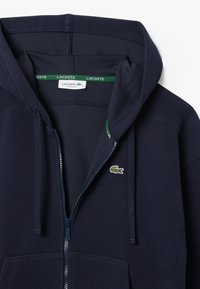 Navy blue zip-up hoodie made of cotton blend. Features a large hood, front pockets, and a green embroidered Lacoste logo on the chest.