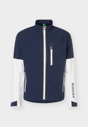 Veste marine et blanche avec fermeture éclair intégrale à l’avant, fermeture éclair de la poche poitrine, deux poches latérales, col montant, et texte « BOGNER » sur la manche droite.