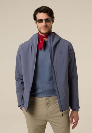 Hombre con gafas de sol, pañuelo rojo al cuello, suéter azul, chaqueta gris y pantalones beige, de pie con una mano en el bolsillo.