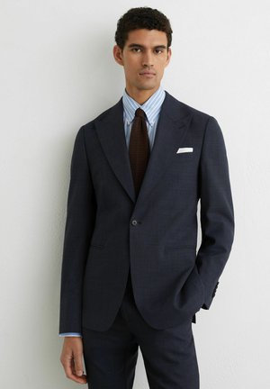 Mladý muž v tmavomodrom obleku s pruhovanou modrou košeľou, hnedou kravatou a bielou vreckovkou stojaci pri bielej stene.