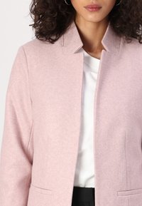 Giacca in misto lana rosa chiaro con colletto a revers, design minimalista e due tasche frontali, indossata su una maglia bianca. Texture morbida.
