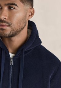 Marineblå lynlåsjakke lavet af blødt fleece stof. Har hætte, snore og en beige foring. Lynlåsen er sølvfarvet.