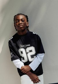 Mustanahi nohik, kellel on punutud juuksed, kannab musta "New York 92" sporditšiki valget pikka varrukat särgi peal, toetudes valgele seinale.