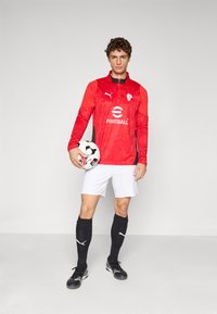 Ung mannlig fotballspiller iført rød trøye, hvite shorts og svarte strømper, som holder en svart-hvit fotball i venstre hånd.