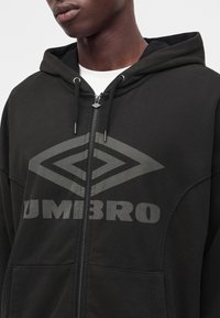 Νέος φοράει μαύρο φούτερ Umbro με φερμουάρ, μεγάλο λογότυπο και κορδόνια, συνδυασμένο με λευκό πουκάμισο από κάτω.