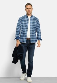 Blau-weiß kariertes Hemd mit Knöpfen, darunter ein weißes T-Shirt, dunkle Jeans, hält eine marineblaue Jacke und weiße Sneaker. Einfaches Design und Passform.