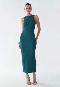 Turkio spalvos suknelė be rankovių, su prigludusia liemene ir plačiu sijonu, pagaminta iš tekstūruotos medžiagos ir vidutinio ilgio pjūvio. Minimalūs akcentai ir švarios linijos.